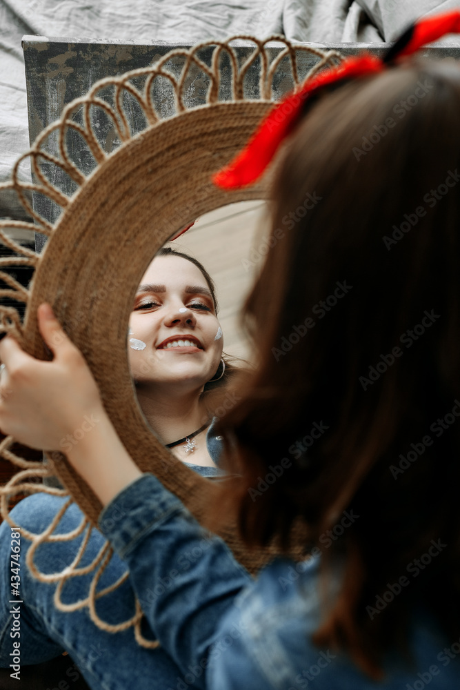 A brunette with a bandage in her hair sits with her back, holding a decorated mirror in her hands and looks at herself in the mirror. She smiles, white streaks of paint on her cheeks. 