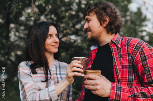 A beautiful, smiling man and a girl in shirts are resting in nature with hot coffee, tea in eco-friendly paper cups, holding in their hands. Photography, concept.