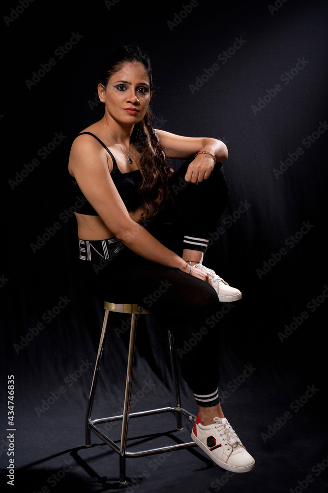 Woman in fitness wear and sneakers sitting on stool. Black background. Looking at camera Woman in fitness wear and sneakers sitting on stool. Black background. Looking at camera