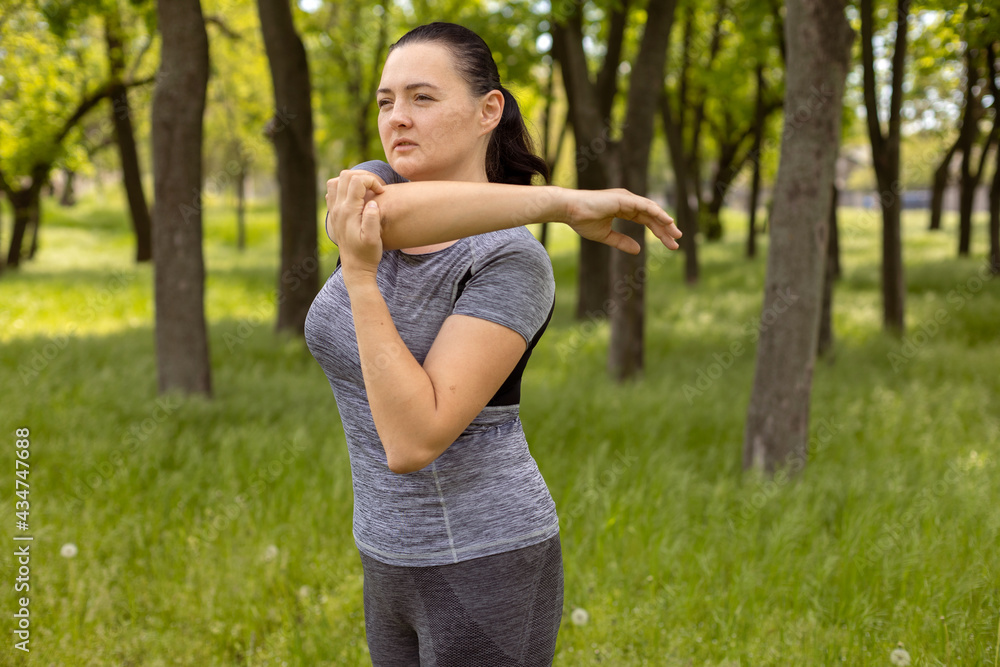 Close up of a young woman warm up her body by stretching her arms to be ready for exercising and do yoga in the park surrounded by nature and warm light afternoon sky. Outdoor workout exercise concept Close up of a young woman warm up her body by stretching her arms to be ready for exercising and do yoga in the park surrounded by nature and warm light afternoon sky. Outdoor workout exercise concept