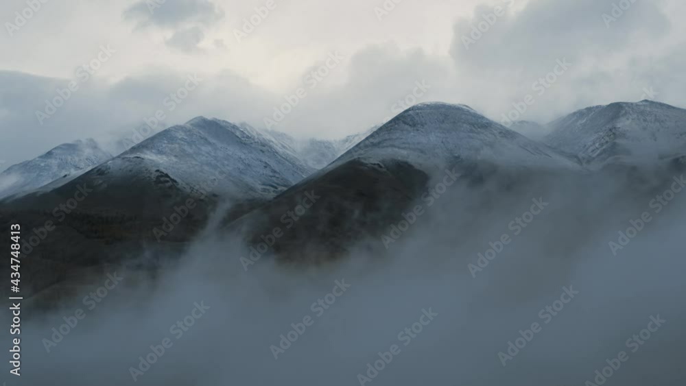 Foggy morning in mountains with snowy peaks. Drone view of Altay mountain range scenery with rocky peaks covered with snow under thick mist in morning Foggy morning in mountains with snowy peaks. Drone view of Altay mountain range scenery with rocky peaks covered with snow under thick mist in morning
