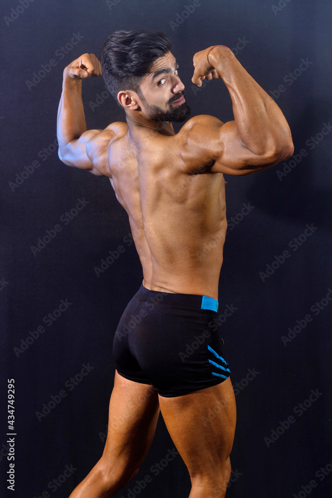 Muscular bodybuilder male in gym. Black background. back profile. Looking at camera. Showing big biceps