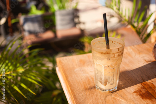 Empty glass with leftovers of coffee latte and straw on a sunlit table in a cozy garden with palm trees and furniture. Hot and sunny tropical morning in a cafe.