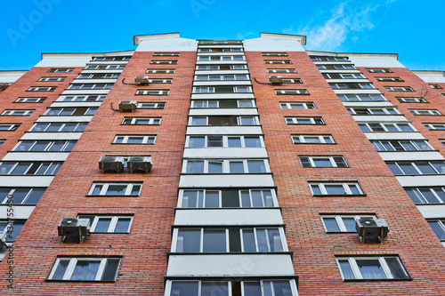 Wall of high modern brick residential building with windows and balconies against blue sky.