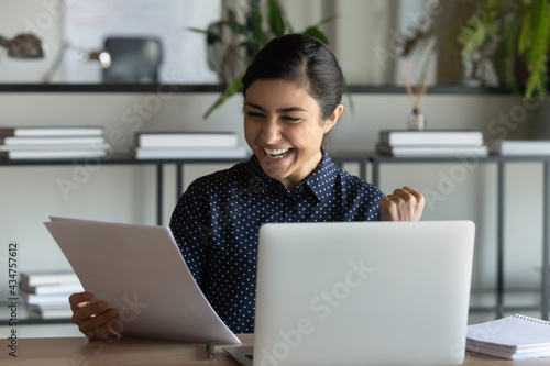 Overjoyed millennial Indian female employee triumph read good news work online on laptop in office. Happy young ethnic woman worker feel euphoric with pleasant hire notice or message in paper letter.