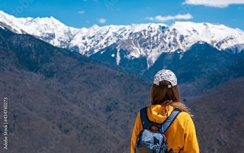 Rear view Young blond woman in yellow hoodie, cap, with backpack on background of snow capped Caucasus mountains. Hiking and travel, active lifestyle, copy space