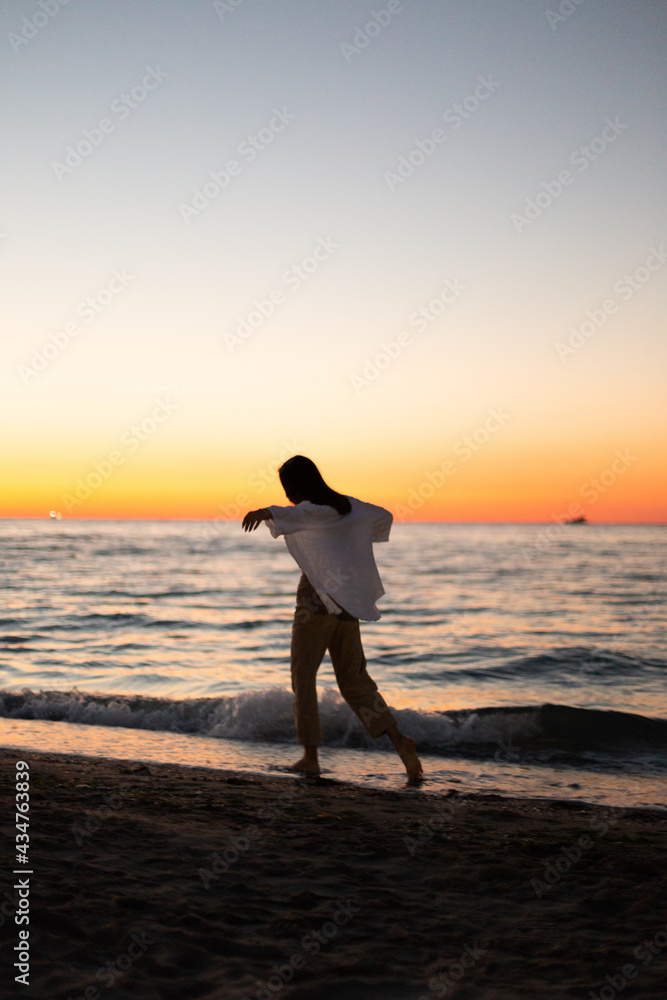 A slender girl in white clothes walks along the beach against the background of the sea. Dawn on the sea. A sportive girl at dawn runs along the embankment of the sea.