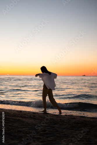 A slender girl in white clothes walks along the beach against the background of the sea. Dawn on the sea. A sportive girl at dawn runs along the embankment of the sea.