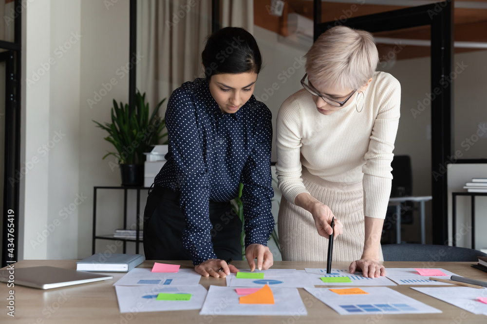 Concentrated multiethnic businesswomen stand at desk in boardroom brainstorm over financial company paperwork together. Focused multiracial female employees discuss startup project at team meeting. Concentrated multiethnic businesswomen stand at desk in boardroom brainstorm over financial company paperwork together. Focused multiracial female employees discuss startup project at team meeting.