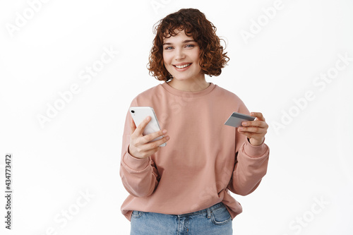 Online shopping. Smiling curly woman holding smartphone and credit card, paying in app, using shop application, order something, standing against white background