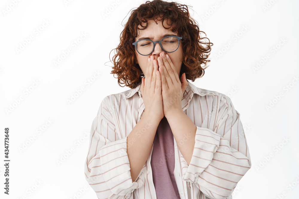 Cute young woman with curly hair and glasses, yawning, covering opened mouth while yawn sleepy, feel tires, sneezing from allergy, standing against white background Cute young woman with curly hair and glasses, yawning, covering opened mouth while yawn sleepy, feel tires, sneezing from allergy, standing against white background