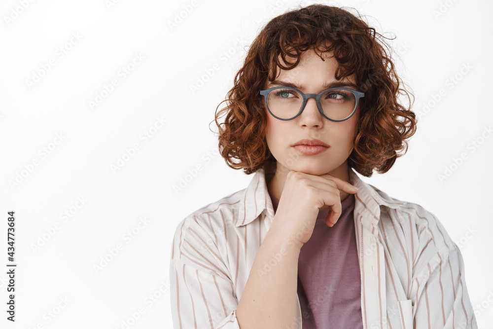 Serious thoughtful girl in glasses, student thinking, looking left at advertisement and pondering, making decision, having a thought, standing over white background Serious thoughtful girl in glasses, student thinking, looking left at advertisement and pondering, making decision, having a thought, standing over white background