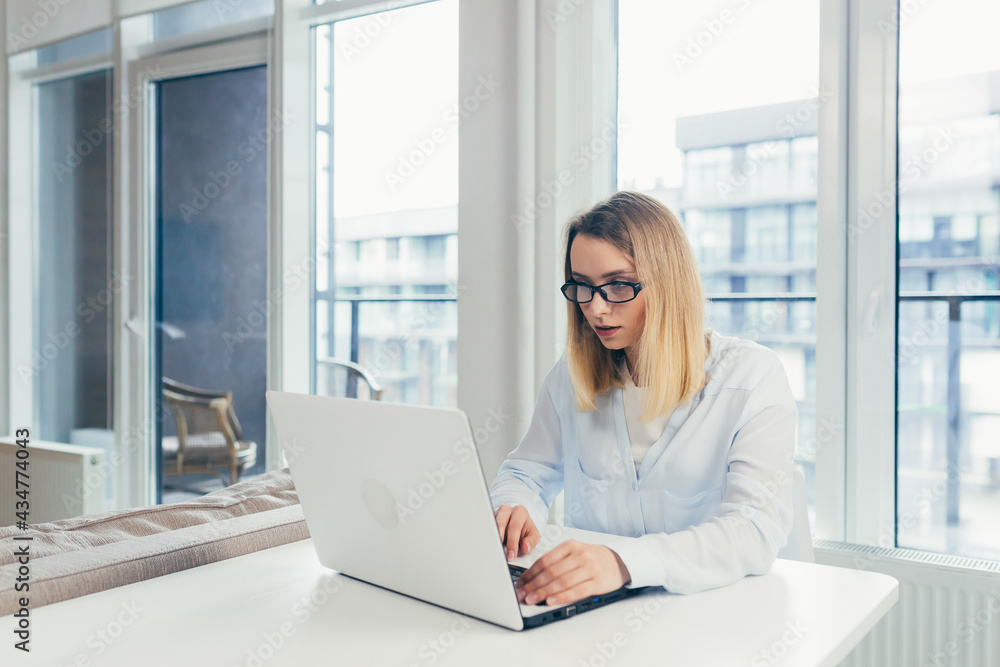 Beautiful blonde woman in casual clothes sitting on a sofa at home, and uses a laptop for video communication with friends and family Beautiful blonde woman in casual clothes sitting on a sofa at home, and uses a laptop for video communication with friends and family
