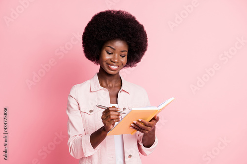 Portrait of pretty cheerful focused wavy-haired girl writing notes story planner isolated on pink pastel color background
