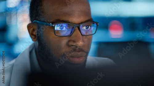 Portrait of Handsome Black Man Wearing Glasses Working Confidently on a Computer. Young Intelligent Male Engineer or Scientist Working in Laboratory. Bokeh Blue Background. Close-up Shot