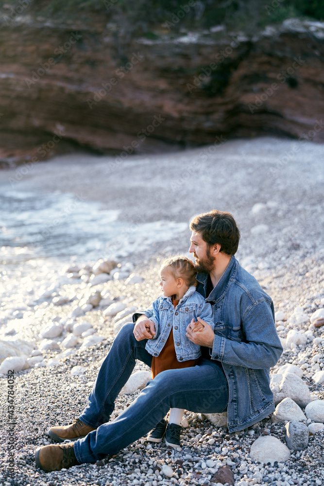 Dad sits on a stone and hugs a little girl on a pebble beach against the backdrop of rocks. Daughter and dad are looking at the sea. Side view