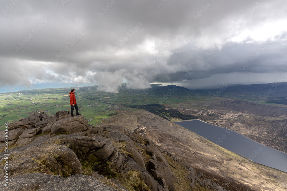 Man in red jacket standing on top of rocky mountain, from behind green fields, Irish Sea, spectacular clouds, Mourne Mountains North Ireland