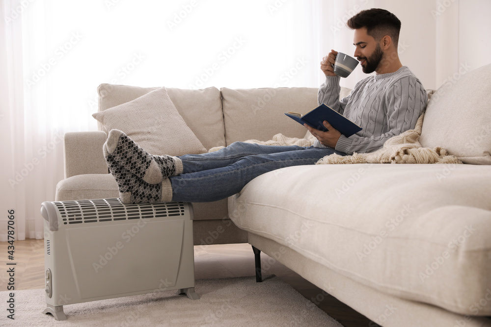 Young man with book and hot drink warming feet on electric heater at home