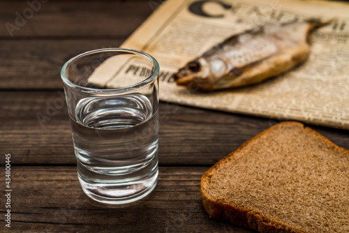 Shot of vodka with a stockfish and newspaper with piece of the black bread on an old wooden table. Close up view