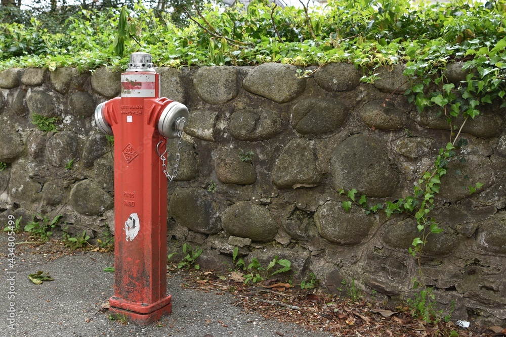 Water hydrant in red color. Fire fighting equipment and water source placed on the street. There is a stone fence on the background. Copy space in the right side. Water hydrant in red color. Fire fighting equipment and water source placed on the street. There is a stone fence on the background. Copy space in the right side.