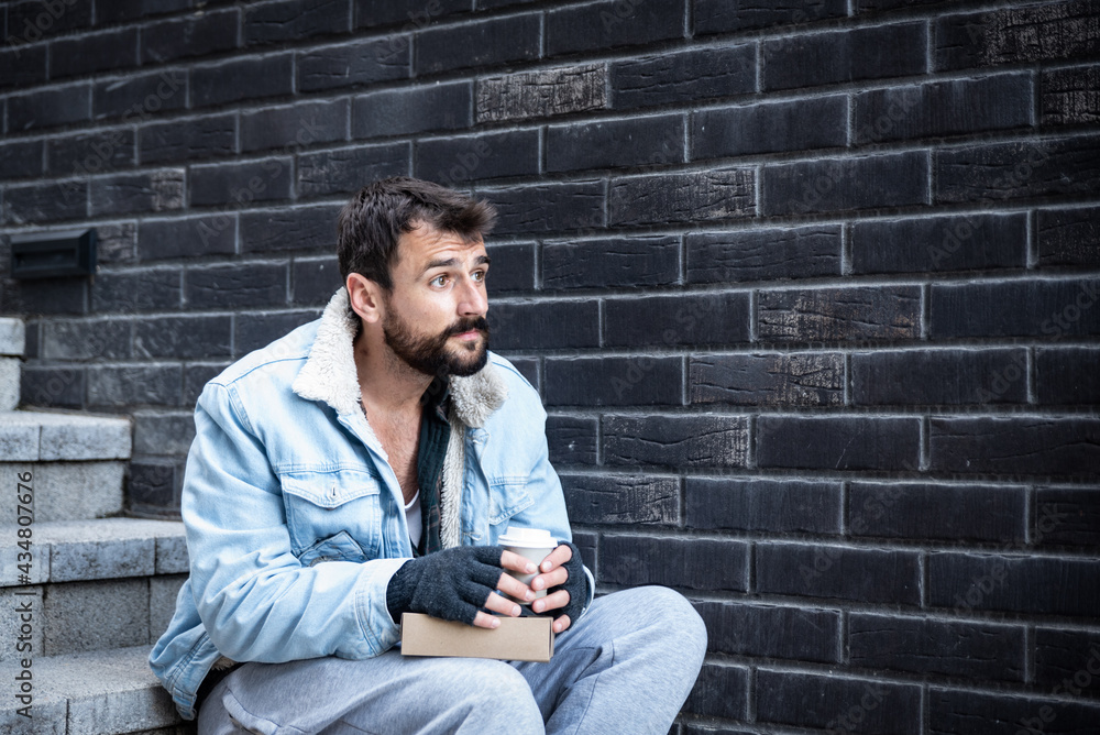 Young confused and desperate homeless man sitting on the staircase drinking coffee and thinking about happy past of his life when he had a job and home, social documentary concept Young confused and desperate homeless man sitting on the staircase drinking coffee and thinking about happy past of his life when he had a job and home, social documentary concept
