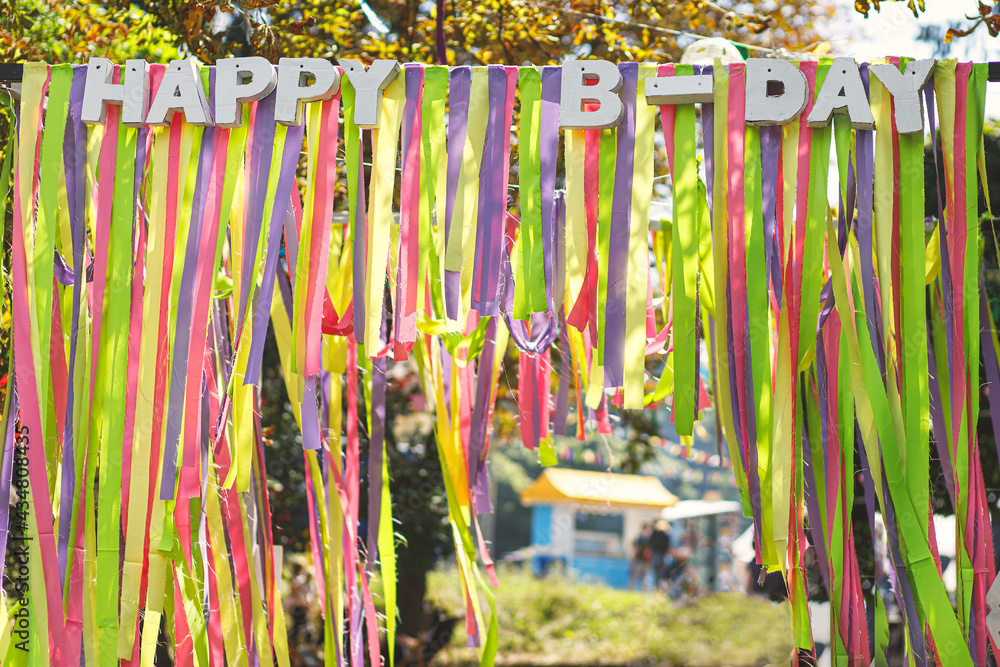 Beautiful colorful ribbons and happy birthday words hanging in sunny park at street food festival, photo zone. Bright rainbow decorations at street food market or birthday. Summer picnic