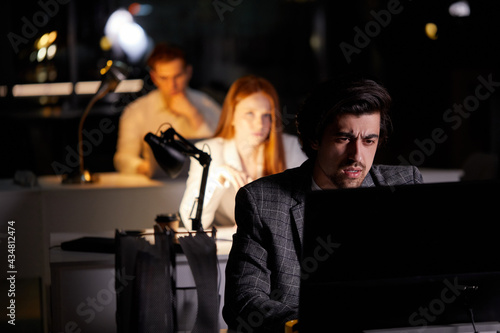 Determined businessman concentrated hard on difficult computer task working late in dark office looking worried, in formal wear, colleagues working on laptop in the background. copy space