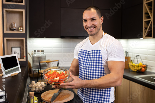 Handsome young man wearing apron holding a bowl of tasty vegetables perfectly cut and sliced