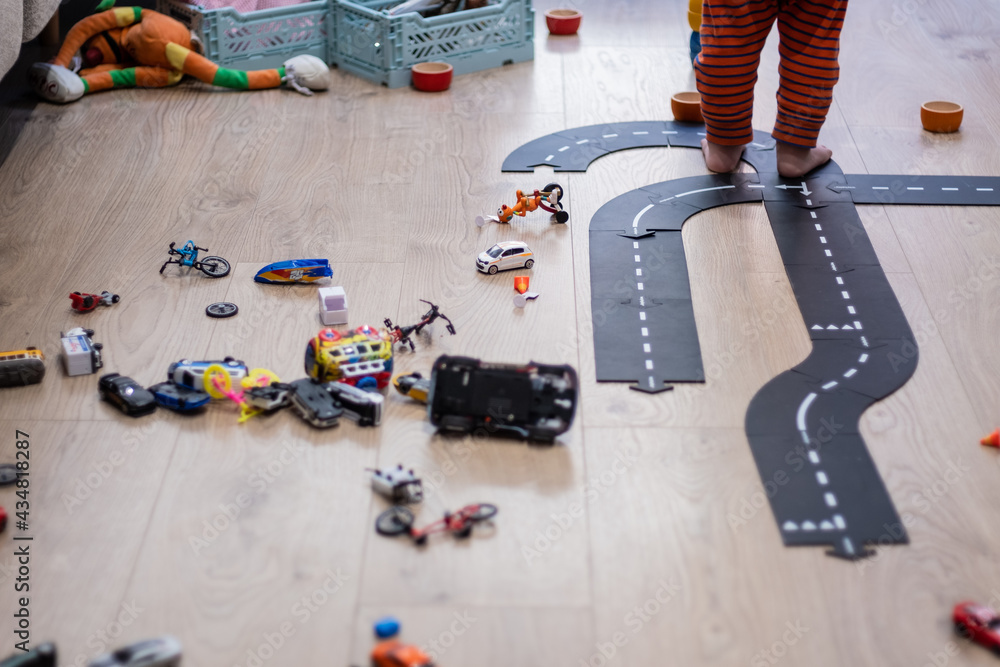 Many toy cars mess and road on a hardwood indoor floor playground with baby legs playing