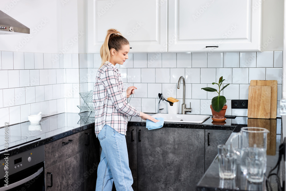 Housewife cleaning the kitchen table and sink with clot blue rag, young female in casual wear housekeeping alone at home, side view Housewife cleaning the kitchen table and sink with clot blue rag, young female in casual wear housekeeping alone at home, side view