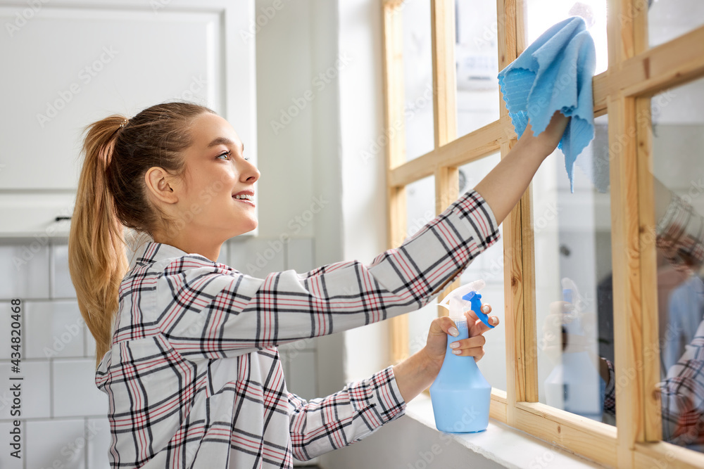 Female cleaning window glass with blue rag. Spring cleanup, housework concept. Side view on smiling woman in checkered shirt Female cleaning window glass with blue rag. Spring cleanup, housework concept. Side view on smiling woman in checkered shirt
