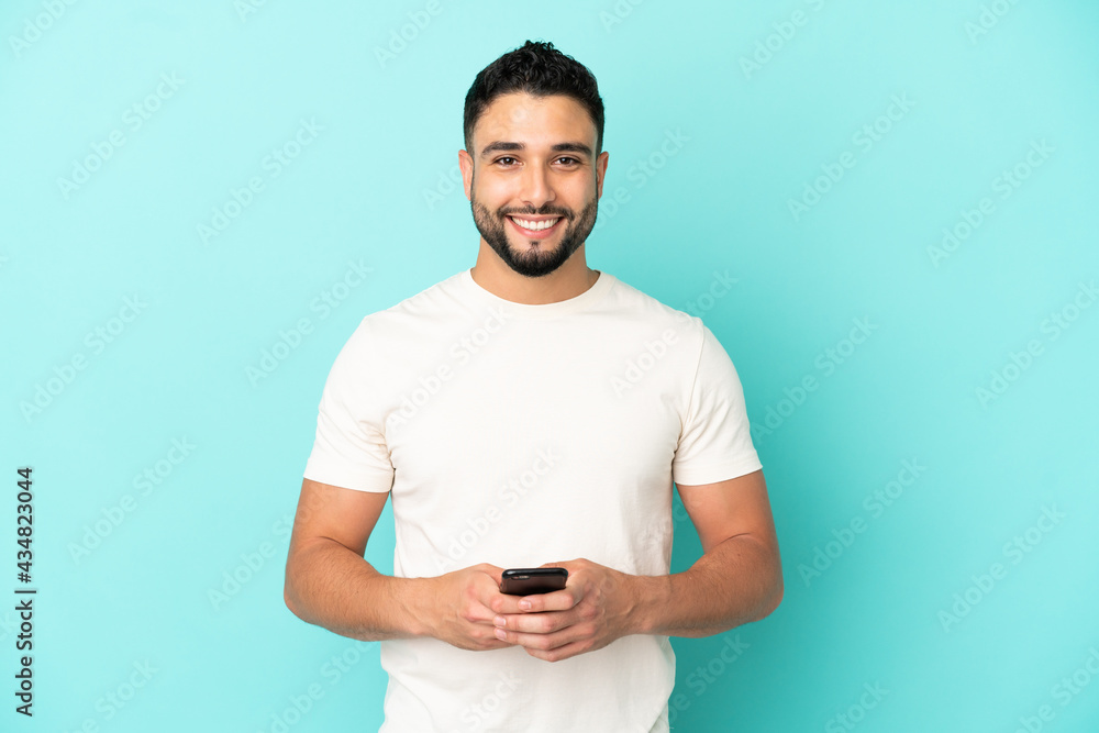 Young arab man isolated on blue background sending a message with the mobile - obrazy, fototapety, plakaty 