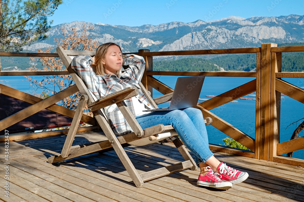 Young woman at the deck of lakehouse, sitting on a wooden chair. Female freelancer working on her laptop sitting outside of the lodge with beautiful lake view. Copy space, close up, background.