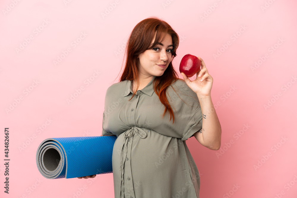 Obraz premium Young redhead caucasian woman isolated on pink background pregnant holding an apple and going to yoga classes
