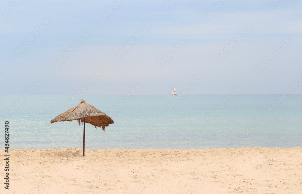 Straw beach umbrella on a tropical sandy beach on a background of blue sea on a sunny day. Idyllic travel and summer vacation concept. Beach umbrella with straw on the sand on a background of blue sea