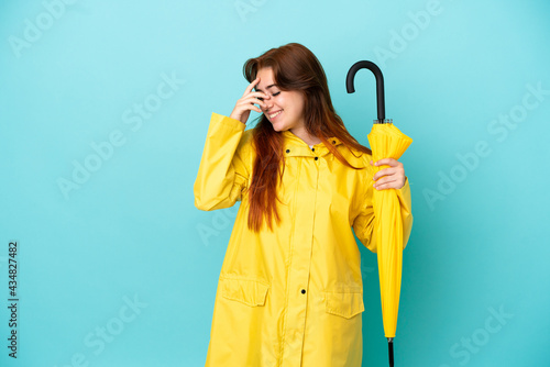 Redhead woman holding an umbrella isolated on blue background laughing