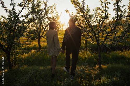 Stylish couple is holding hands and walking in the spring blooming garden at sunset.