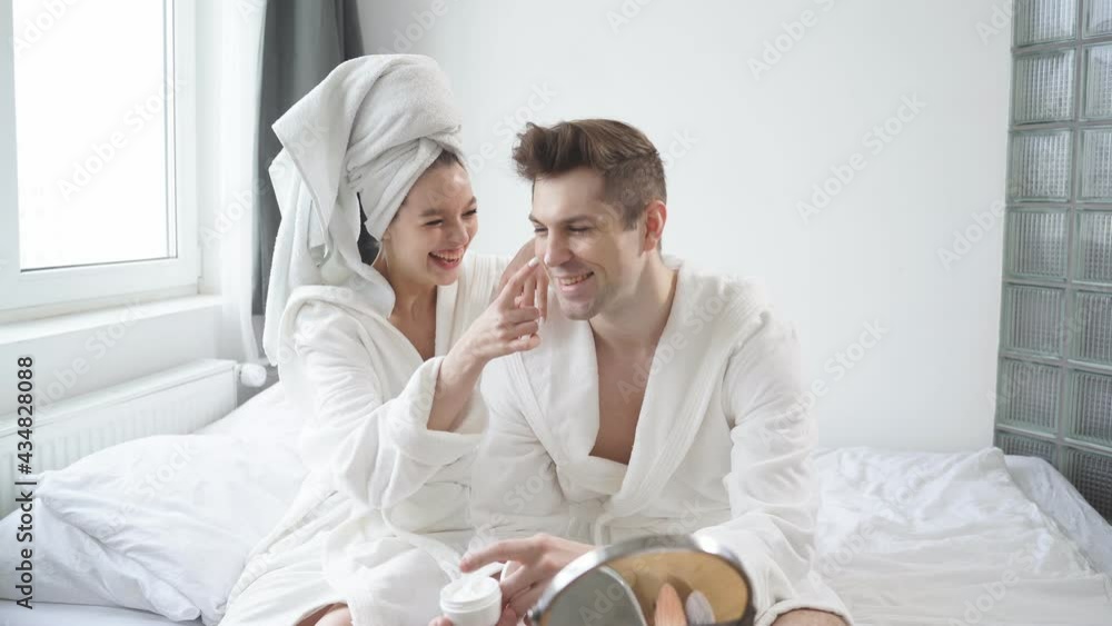 Morning preparing of beautiful caucasian couple in bathrobe, use moisturizing cream sitting in bright modern bedroom, wearing bathrobe and towel Morning preparing of beautiful caucasian couple in bathrobe, use moisturizing cream sitting in bright modern bedroom, wearing bathrobe and towel