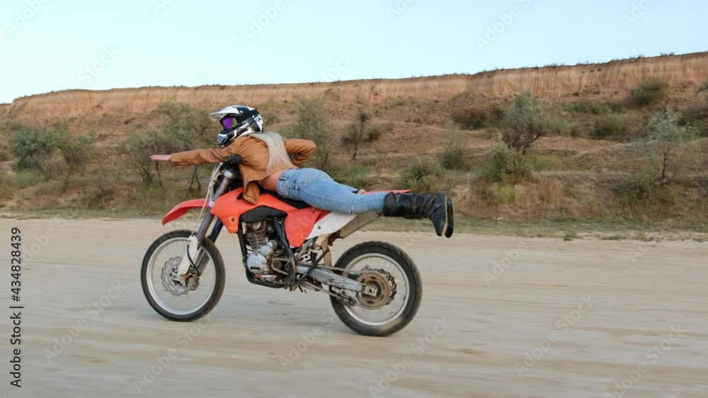 Brave young woman does stunts riding red motorbike on ground road in countryside area against dry grass and thickets field Brave young woman does stunts riding red motorbike on ground road in countryside area against dry grass and thickets field