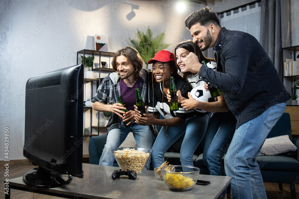 Four multiracial fans emotional cheering for football team during match. Happy friends watching sport on television. Young people screaming and gesturing.