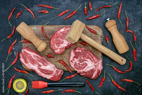 Beef Steaks for Grilling Or Frying, Overhead View. Marbled Raw Loin Beef Steaks, Wooden Hammer, Salt and Pepper Mill On Cutting Board. Raw Striploin Marbled Beef Steaks on Black Background, Top View.