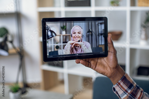 Close up of male hand holding digital tablet with muslim woman in hijab on screen. Young african man talking with girlfriend through video call during pandemic.