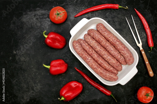 Sausages For BBQ Grilling or Frying in White Ceramic Dish And Red Peppers on Grungy Black Background, Top View. Raw Beef or Lamb Barbecue Sausages in Natural Casing in White Tray, Overhead View.