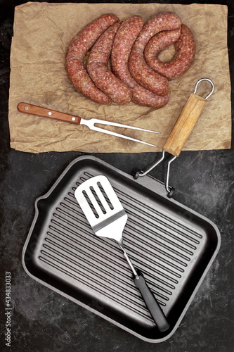 Raw Stuffed Sausages, Empty Grill Pan and Grill Tools On Rustic Black Table Background, Top View. Raw Sausages In Natural Casing. Sausages For Grilling or Frying On Paper Overhead View.