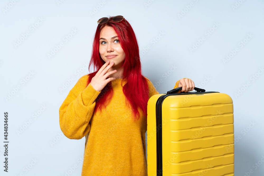 Traveler girl holding a suitcase isolated on blue background scheming something Traveler girl holding a suitcase isolated on blue background scheming something