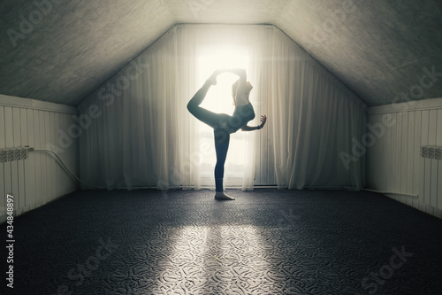 Young female stretching doing morning yoga exercises near big window at home