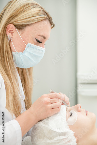 Cosmetologist wearing protective face mask lengthening female lashes in beauty salon