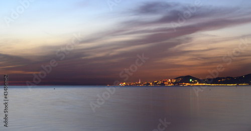 Plain water surface of the Black Sea. Dark clouds are colored with the set sun. Lights of the city Adler in the background. The beauty of sea sunsets.
