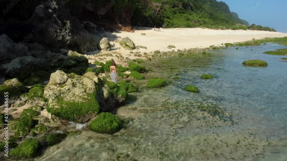 Pretty woman walks in the beach near to rocks with green seaweed around big cliff in Nunggalan Beach, Bali, Indonesia. High quality 4k aerial footage Pretty woman walks in the beach near to rocks with green seaweed around big cliff in Nunggalan Beach, Bali, Indonesia. High quality 4k aerial footage