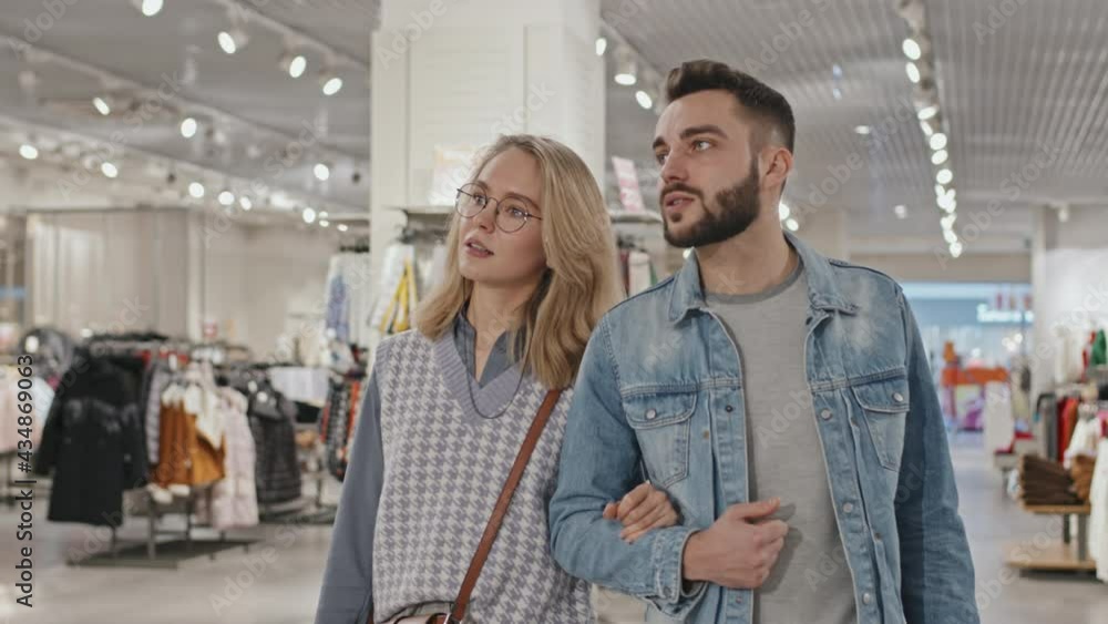 Medium shot of stylish young Caucasian couple talking while walking towards camera shopping in clothing store together Medium shot of stylish young Caucasian couple talking while walking towards camera shopping in clothing store together